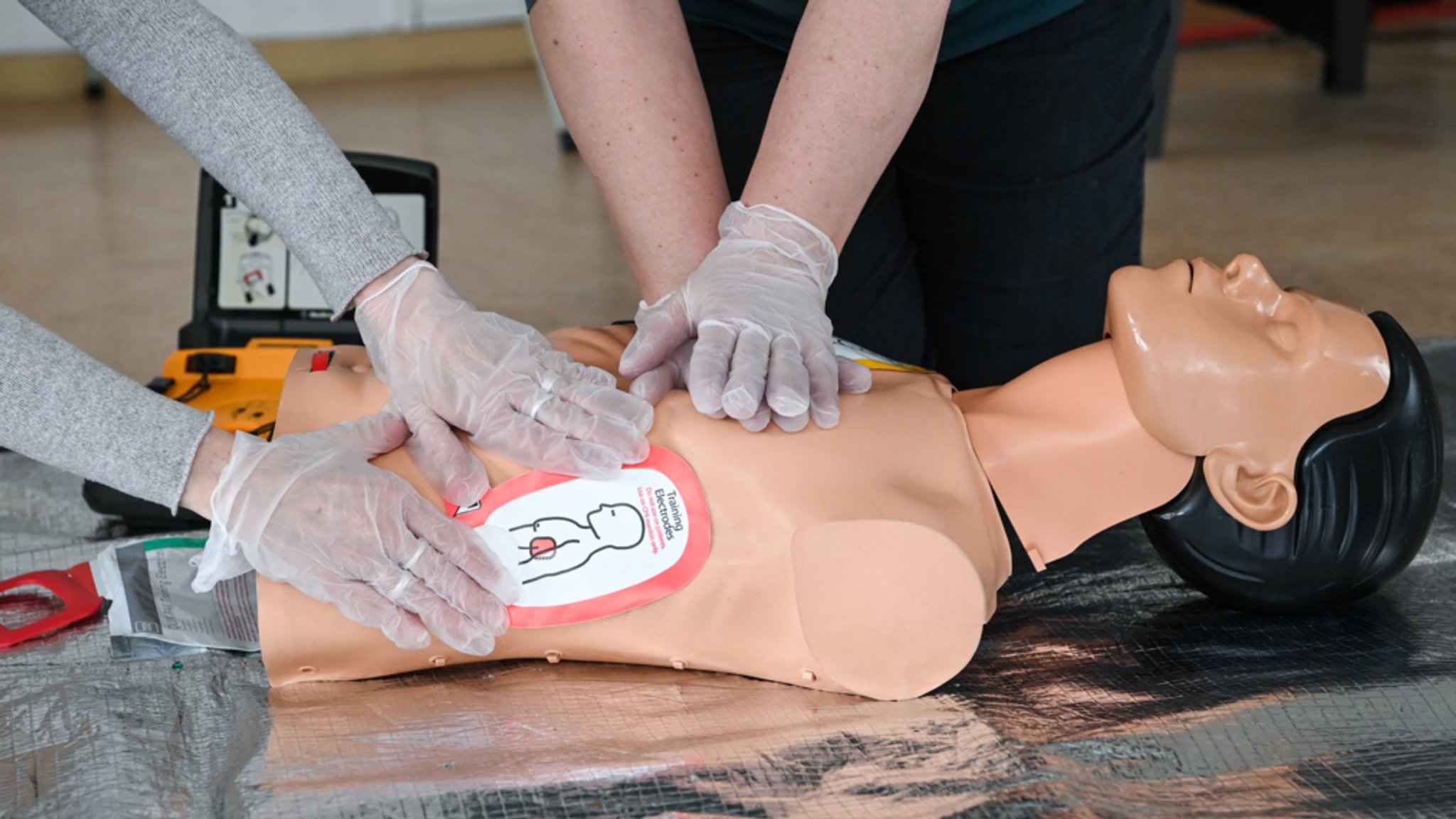 Die Hände zweier Menschen in Handschuhen üben Herzdruckmassage an einer Übungspuppe für Erste Hilfe.