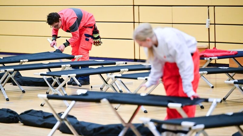 03.01.2026, Berlin: Ehrenamtliche Mitarbeiter des DRK bauen während eines Stromausfalls im Südwesten Berlins in einer Notunterkunft Feldbetten auf. Vom Brand einer Kabelbrücke sind 50.000 Haushalte und 2.000 Gewerbebetriebe betroffen. Foto: Sebastian Christoph Gollnow/dpa +++ dpa-Bildfunk +++ | Bild: dpa-Bildfunk/Sebastian Gollnow 03.01.2026, Berlin: Ehrenamtliche Mitarbeiter des DRK bauen während eines Stromausfalls im Südwesten Berlins in einer Notunterkunft Feldbetten auf. Vom Brand einer Kabelbrücke sind 50.000 Haushalte und 2.000 Gewerbebetriebe betroffen. Foto: Sebastian Christoph Gollnow/dpa +++ dpa-Bildfunk +++