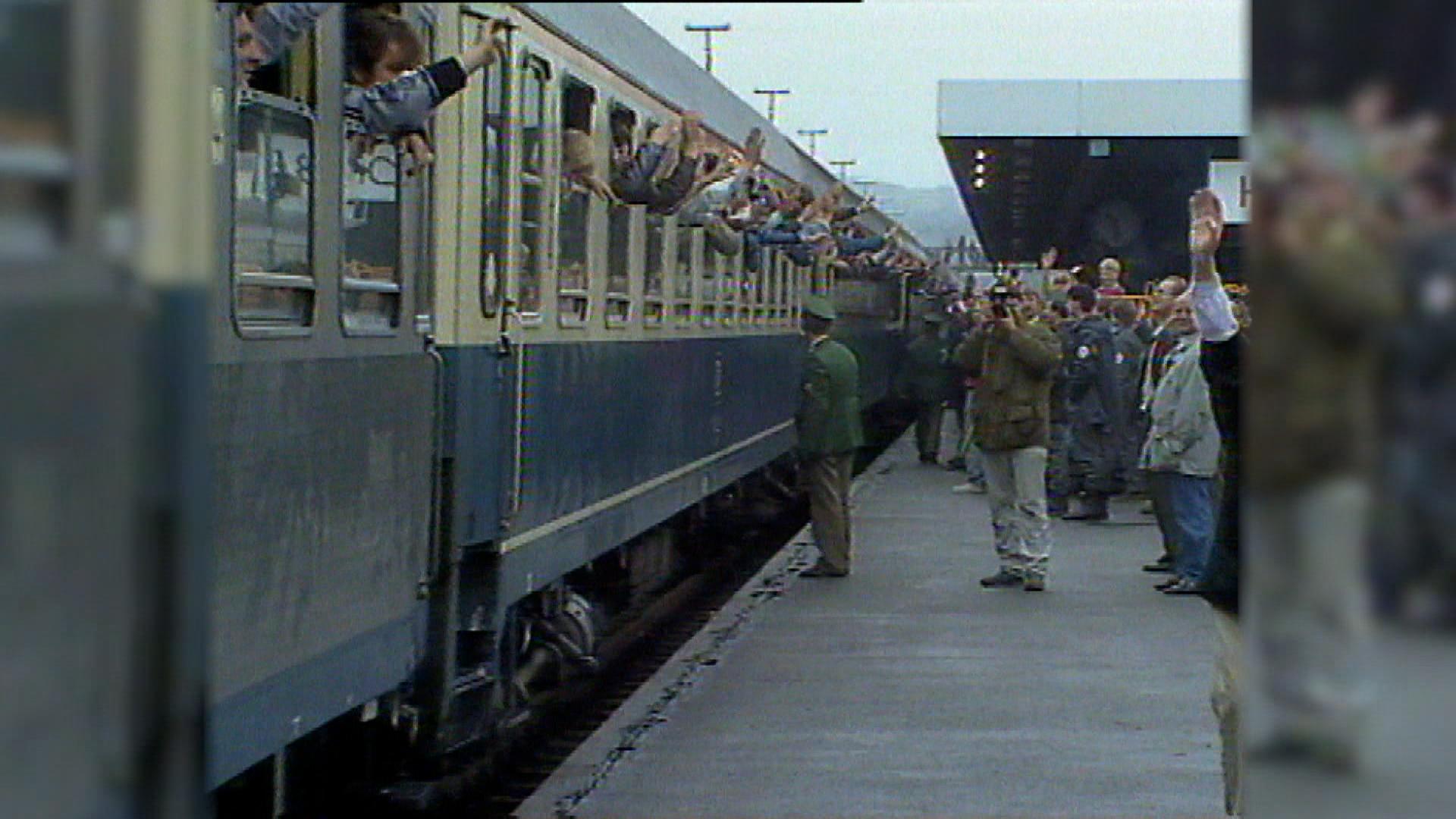 Menschen winken aus einem Zug, am Bahnsteig stehen viele Menschen und Polizisten. 