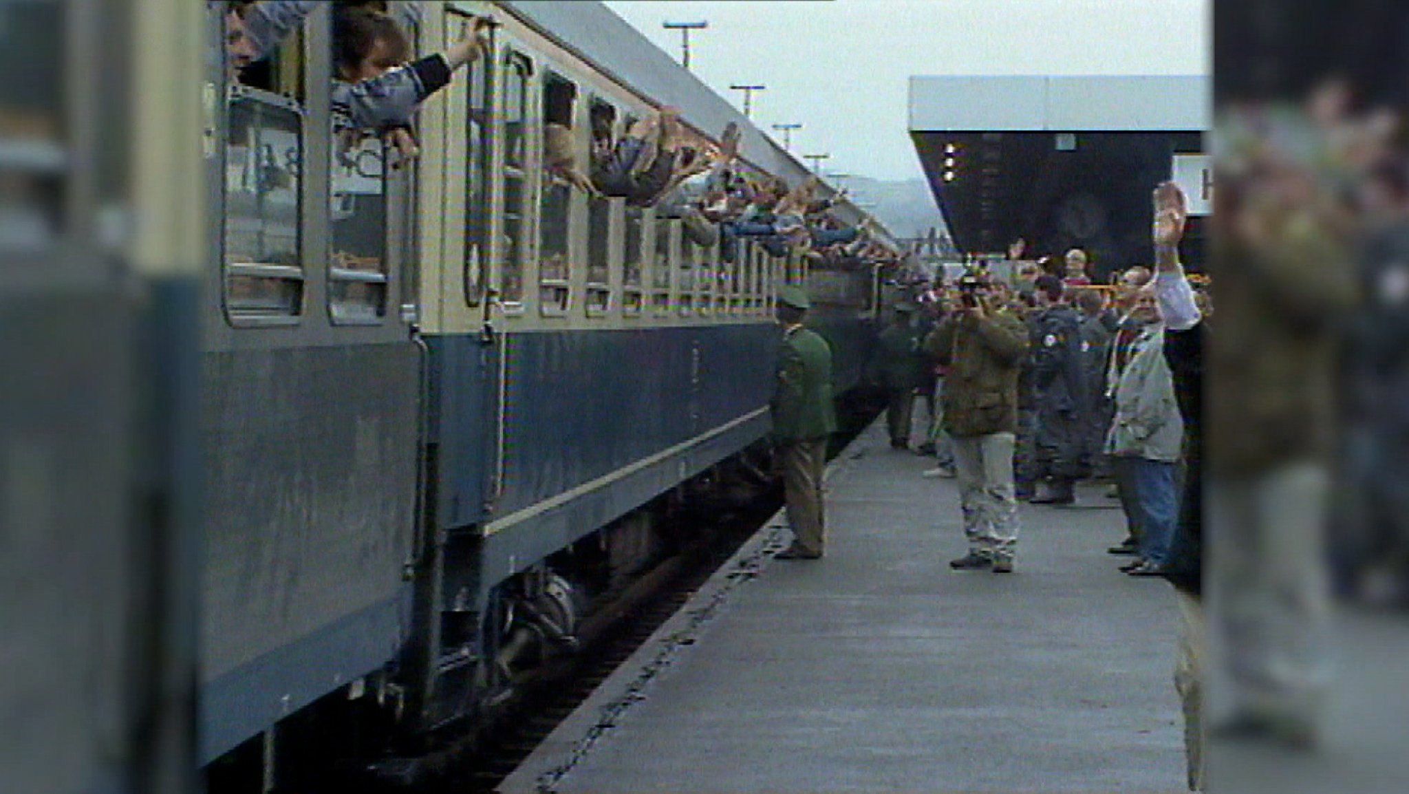 Menschen winken aus einem Zug, am Bahnsteig stehen viele Menschen und Polizisten. 