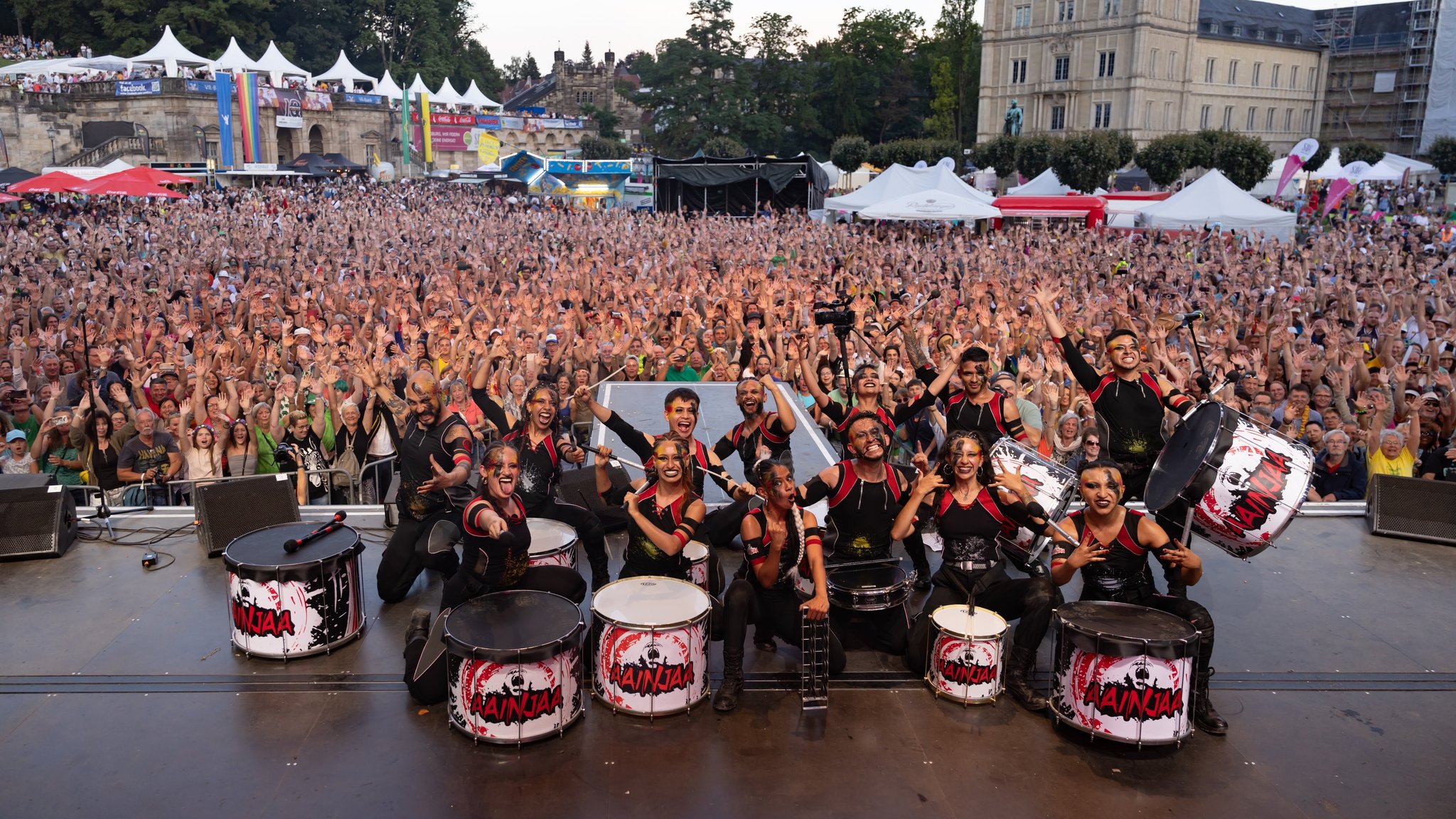 Eine Trommlergruppe steht auf eine Bühne und posiert mit ihren Trommeln für das Foto. Im Hintergrund ist ein mit Menschen gefüllter Platz zu sehen, die alle die Arme in die Höhe reißen. 