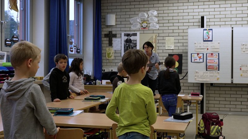 Schulkinder stehen mit einer Lehrerin in einem Klassenzimmer.