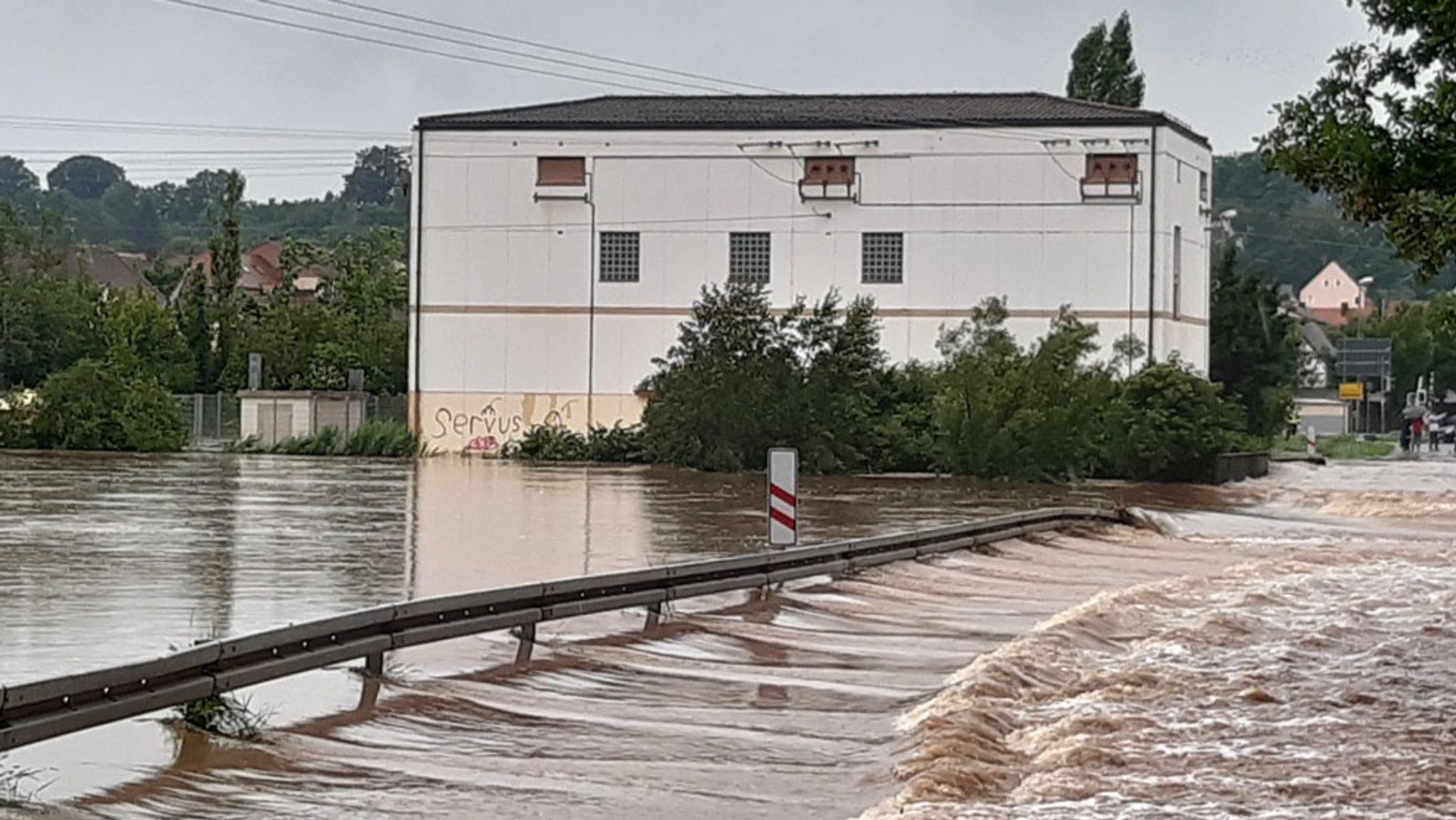 Wasser spült über einer Straß, im Hintergrund ein breites Trafohaus