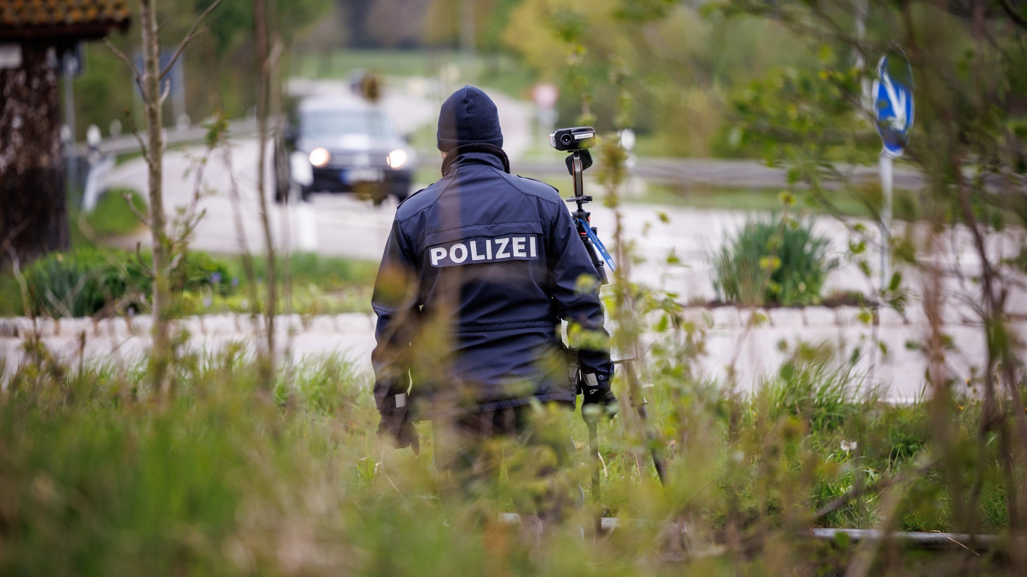 Ein Polizist steht versteckt am Straßenrand einer Staatsstraße außerorts bei Wessobrunn (Bayern) neben einer aufgebauten Laserpistole zur Geschwindigkeitsmessung.