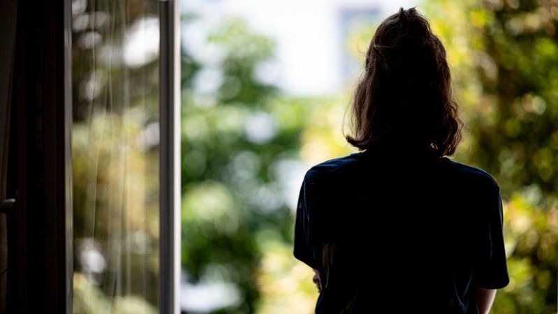Symbolbild Einsamkeit: Eine junge Frau steht in ihrer Wohnung an einem Fenster.