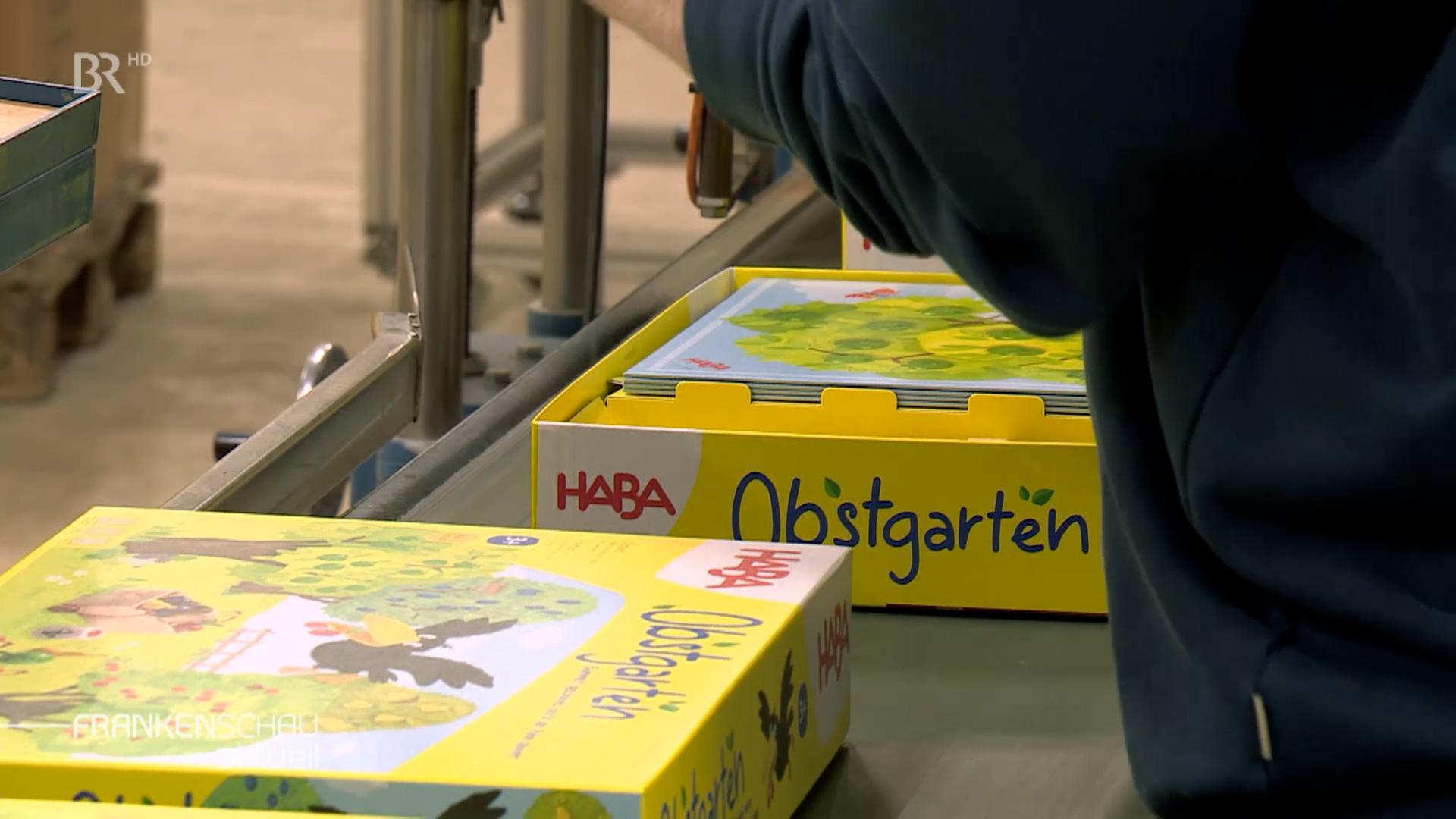 Spiele der Marke Haba auf einem Förderband in einer Halle. 