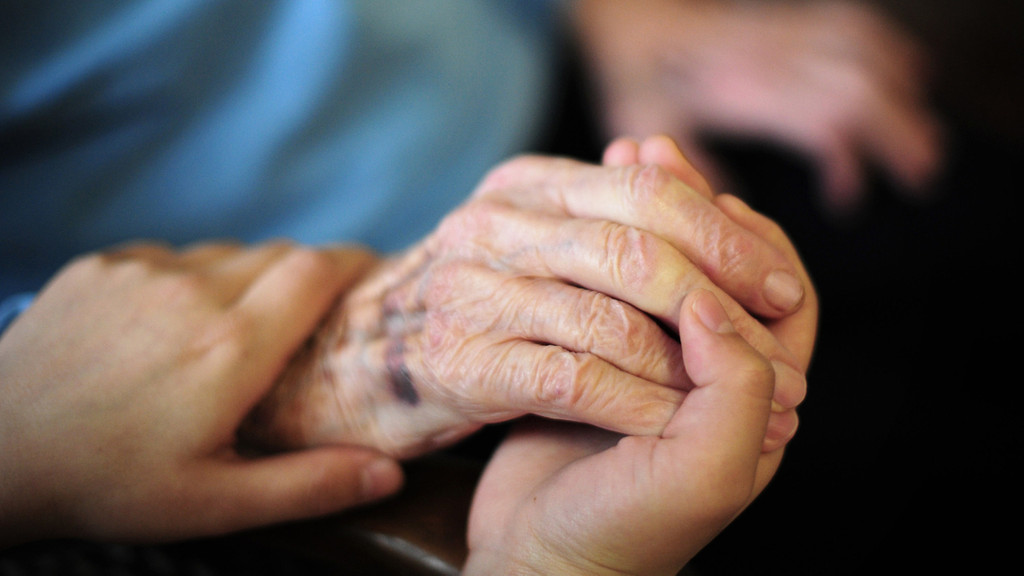 Hand einer Pflegerin hält eine Hand eines älteren Menschen.