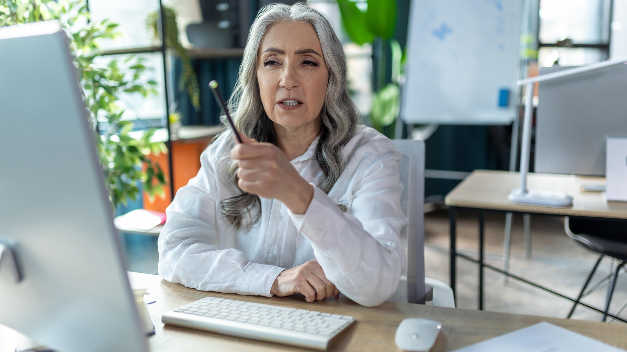 Eine Frau mit grauen, langen Haaren sitzt an einem Schreibtisch vor einem PC. Beschwerden während der Wechseljahre beeinträchtigen viele Frauen auch am Arbeitsplatz.
