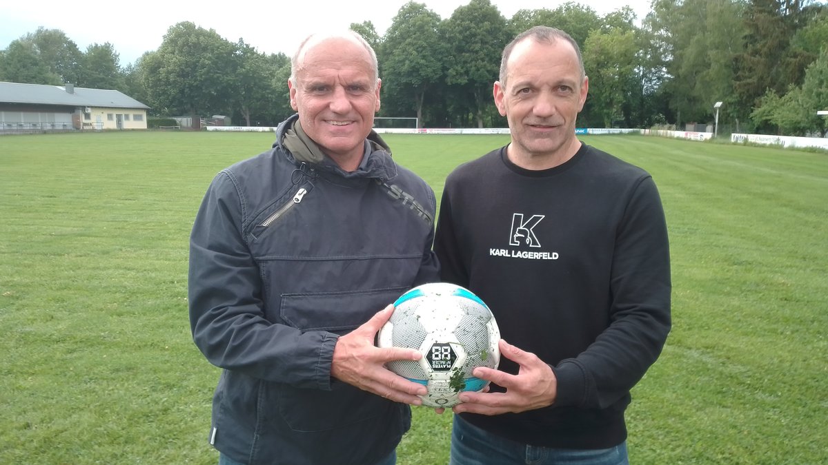 Martin Schneider und Pero Škorić stehen auf einem Fußballfeld und halten gemeinsam einen Ball in die Kamera. 