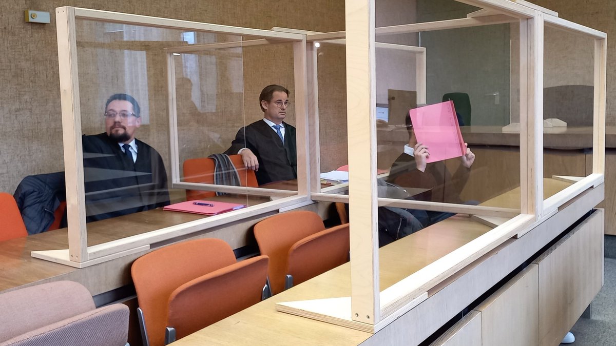 Der Angeklagte am 17.2.23 im Gerichtssaal, vor dem Gesicht eine rosafarbene Mappe. In der Reihe hinter ihm seine zwei Anwälte. Auf den Tischen stehen Gestelle mit Plexiglasscheiben.