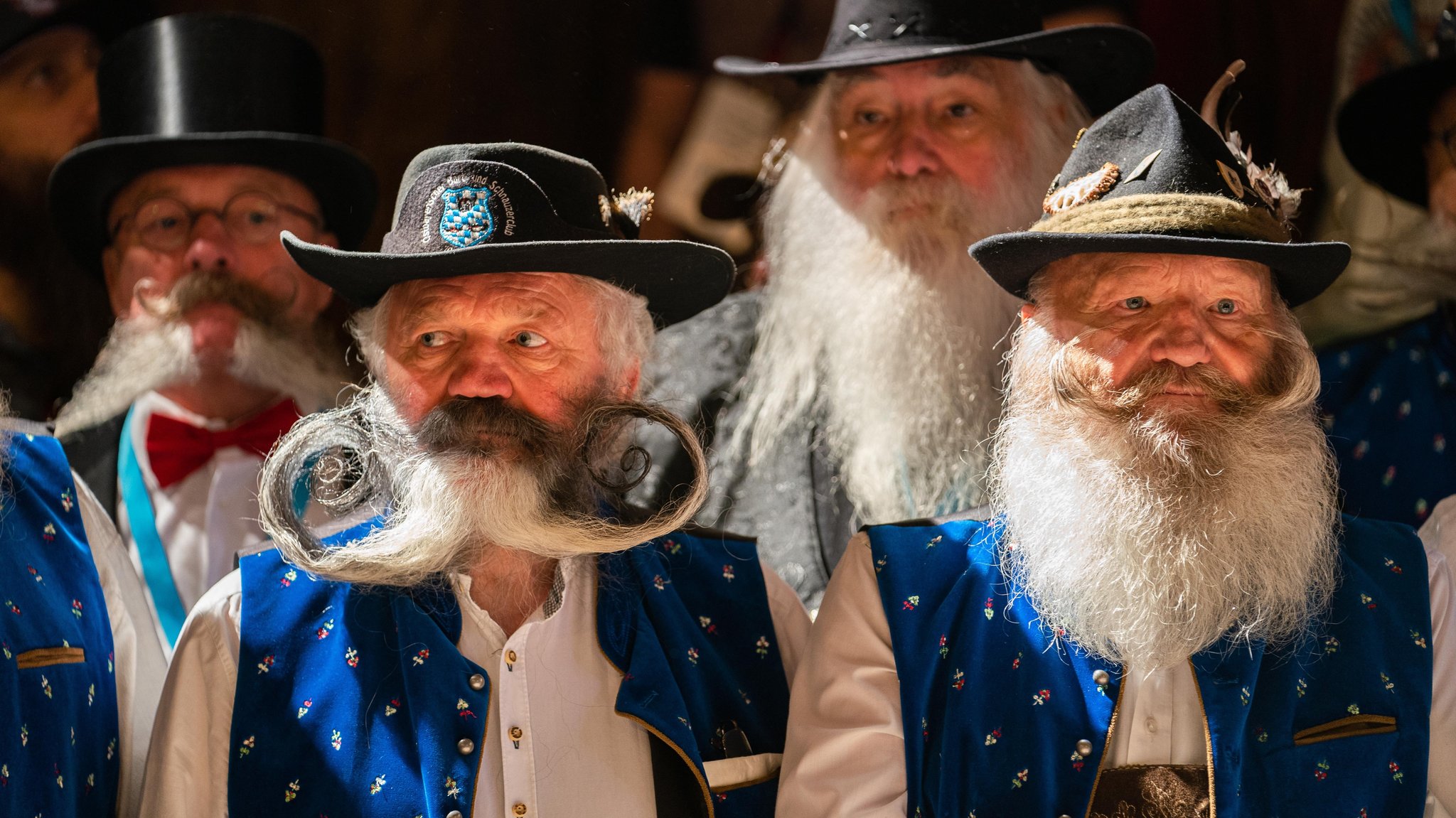 Im Vordergrund sind zwei ältere Herren in blauer Tracht und weißem Hemd zu sehen. Sie tragen Hut und haben beide lange weiße Bärte. Der linke hat seinen gesamten Bart in zwei Hälften zu beiden Seiten seines Gesichts gezwirbelt. Der Rechte trägt auch Vollbart und hat seinen Schnurrbart ebenfalls zu beiden Seiten gezwirbelt. 