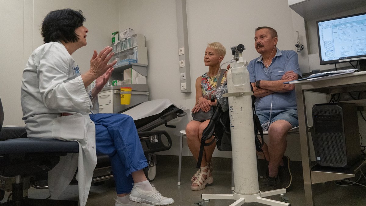 In einem Behandlungszimmer sitzen drei Menschen: Eine Frau mit weißem Kittel, ihr gegenüber ein Ehepaar aus Frau und Mann. Der Mann wird über einen Schlauch mit Sauerstoff aus einer Gasflasche versorgt.