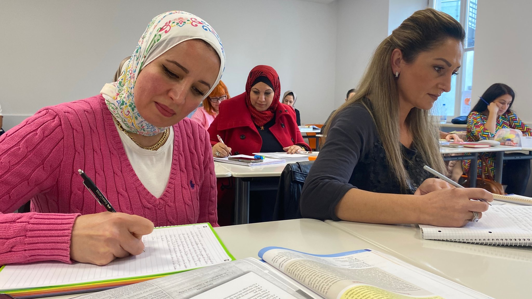 Sechs Frauen in einem Klassenzimmer, die etwas schreiben. Drei tragen ein Kopftuch, drei nicht.