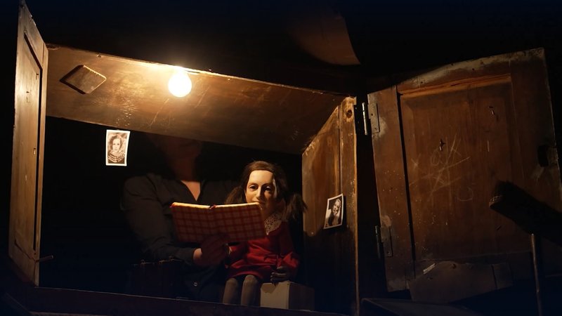 Eine Puppe, die Anne Frank darstellen soll, sitzt in einem roten Kleid auf einer Holzkiste, ihr Tagebuch auf dem Schoß. | Bild: BR/Christian Akber-Sade Eine Puppe, die Anne Frank darstellen soll, sitzt in einem roten Kleid auf einer Holzkiste, ihr Tagebuch auf dem Schoß.