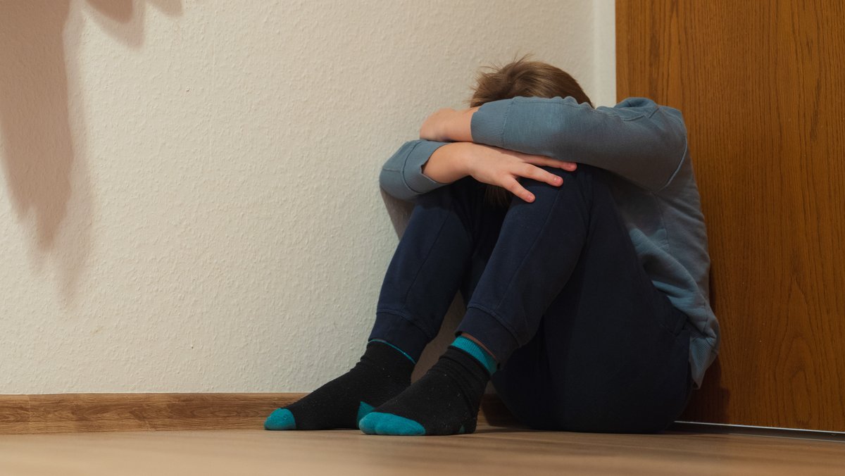 Ein Junge kauert sich in der Ecke eines Zimmers am Fußboden sitzend zusammen. (Symbolbild)