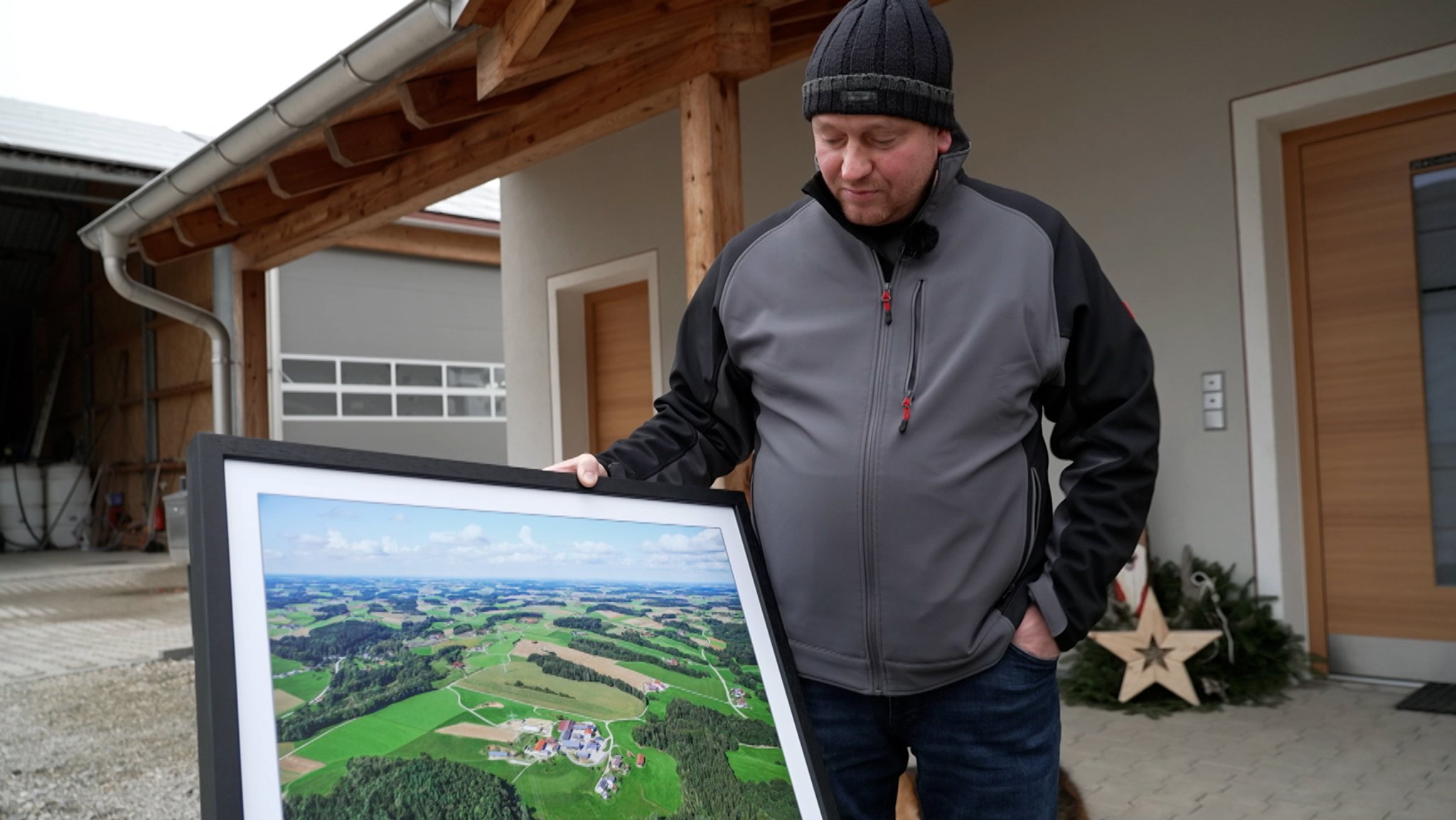 Der Landwirt Stefan Hirler hat eine große, gerahmte Luftaufnahme in der Hand, auf der sein Hof abgebildet ist. Er sieht auf das Bild und steht vor seinem Hauseingang.