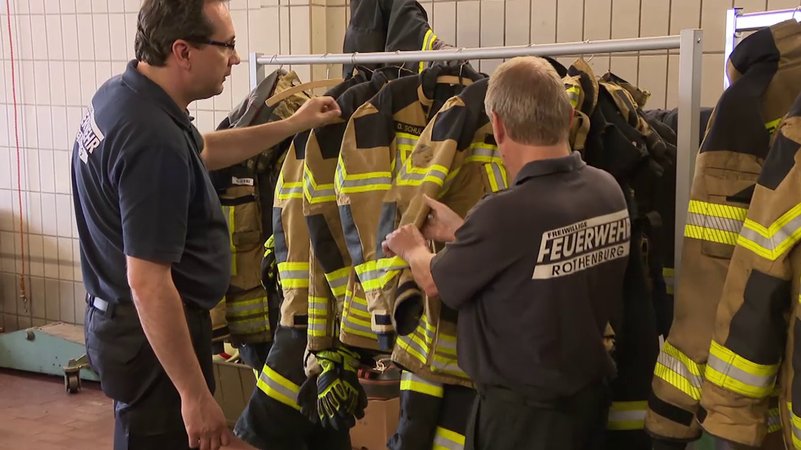 Zwei Männer der Feuerwehr überprüfen die Einsatzkleidung, die an Kleiderbügeln hängt.