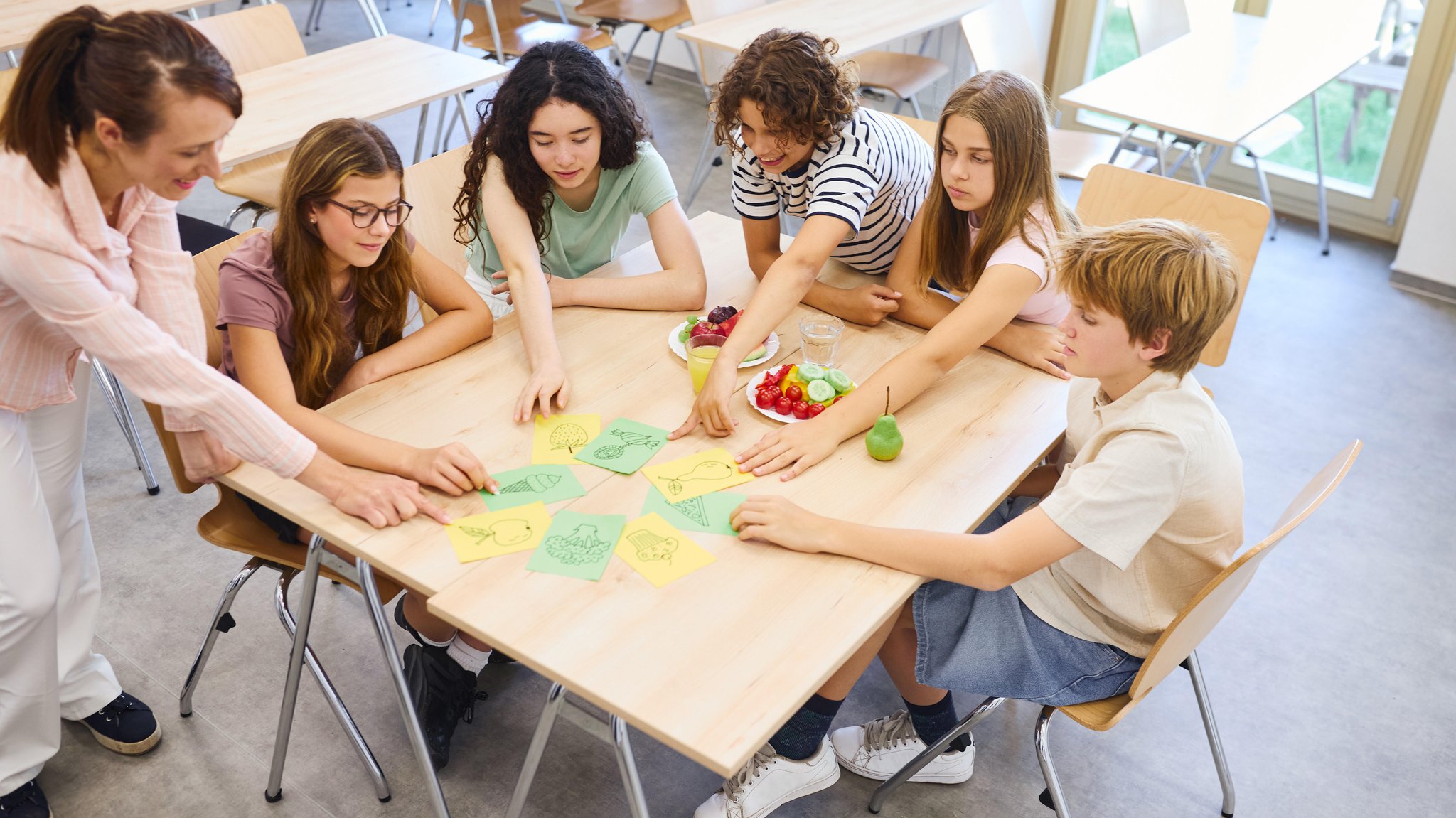 (Symbolbild) Schüler sitzen rund um einen Tisch, eine Lehrerin steht daneben und deutet auf eine der Karten, die auf dem Tisch liegen. | Bild: picture alliance / Zoonar / Robert Kneschke (Symbolbild) Schüler sitzen rund um einen Tisch, eine Lehrerin steht daneben und deutet auf eine der Karten, die auf dem Tisch liegen.