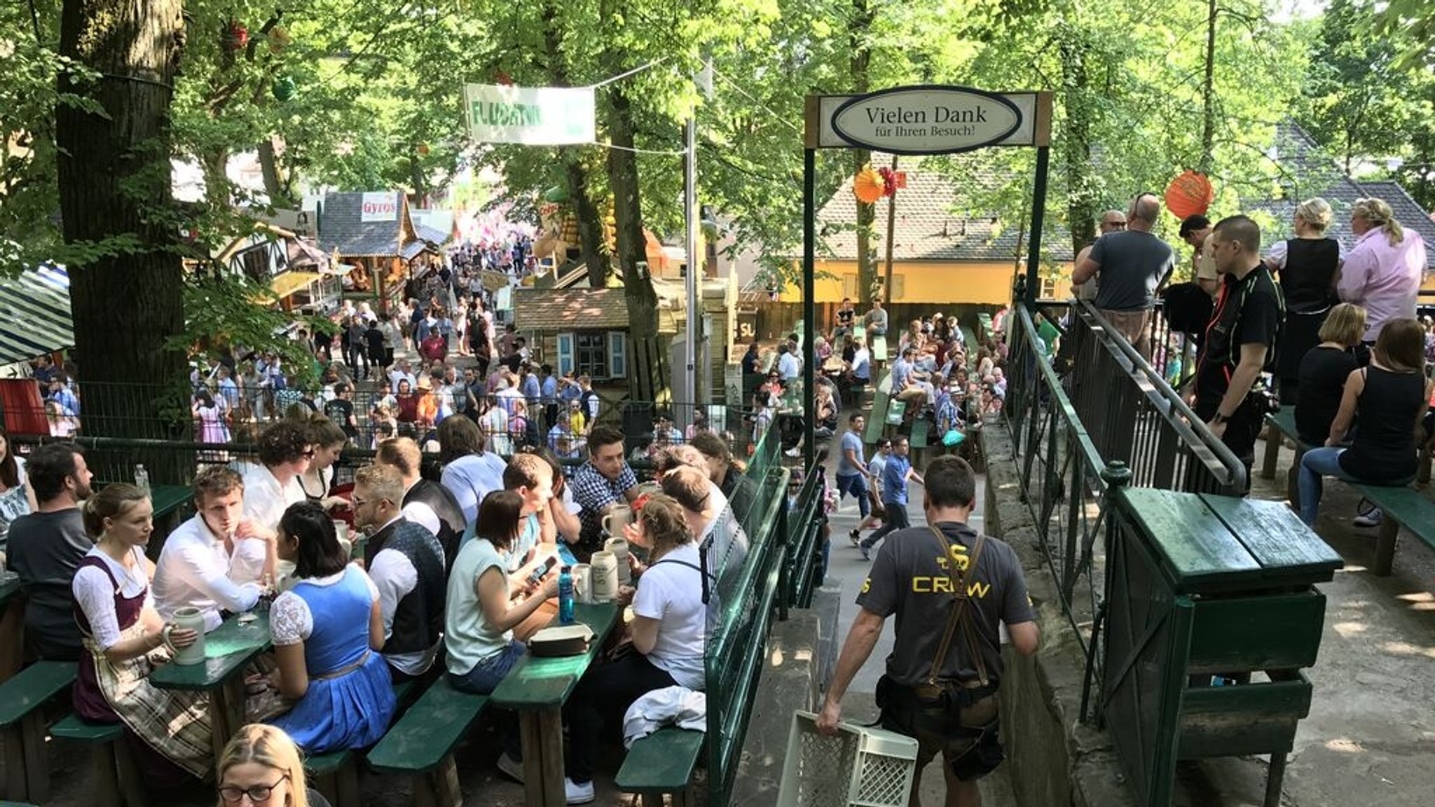Besucher der Erlanger Bergkirchweih sitzen an Bierbänken unter den schattigen Bäumen und trinken Bier.