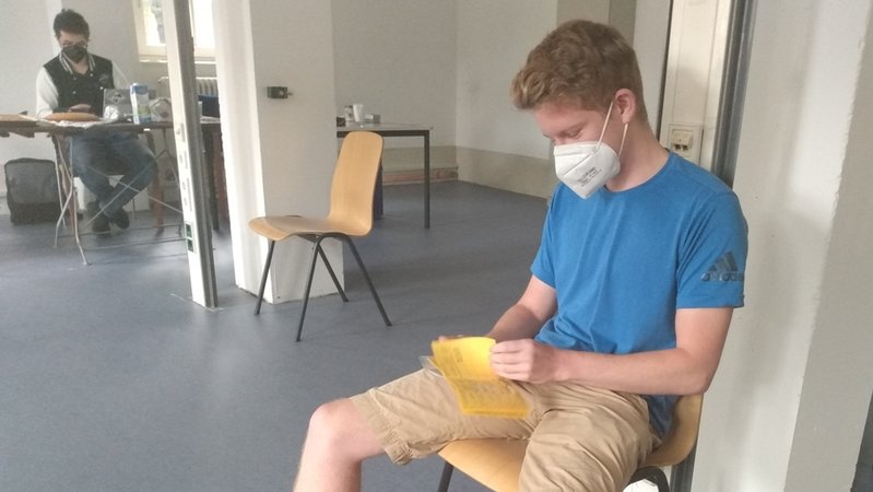 Student sitzt auf einem Stuhl und blättert in seinem gelben Impfpass