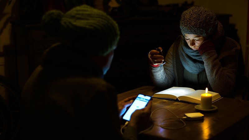 (Symbolbild) Zwei Personen im Dunkeln, eine liest im Kerzenschein, die andere lädt ihr Handy an einer Powerbank auf | Bild: picture alliance / Shotshop | °ts (Symbolbild) Zwei Personen im Dunkeln, eine liest im Kerzenschein, die andere lädt ihr Handy an einer Powerbank auf