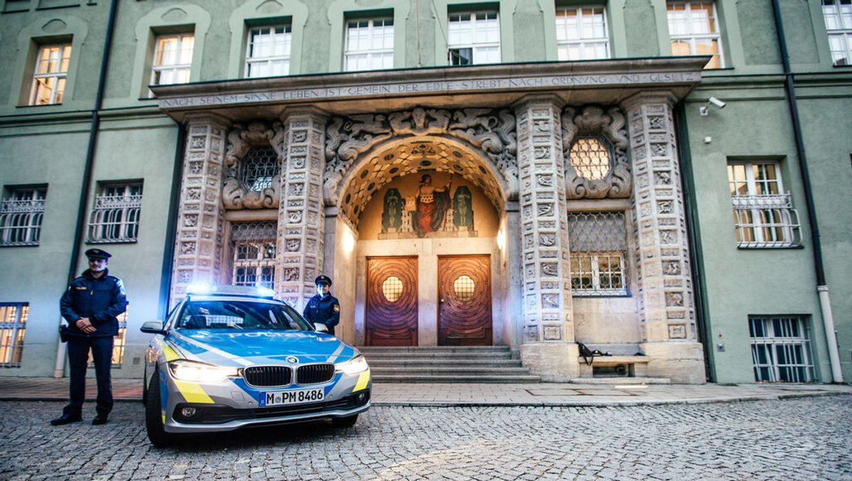 Eine Polizistin und ein Polizist stehen vor dem Eingang des Polizeipräsidiums München vor einem Polizeiauto mit Blaulicht.