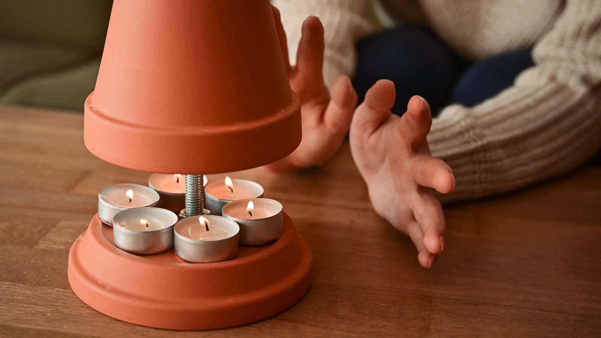 Eine Person wärmt sich symbolisch an einem selbstgebauten Teelichtofen 