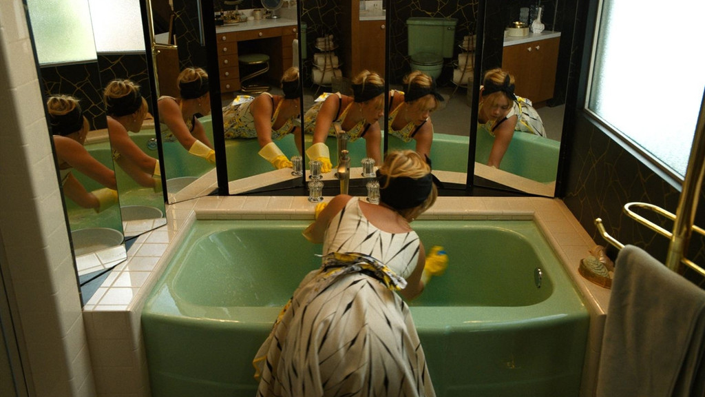 Vexierspiel im Badezimmer: Alice im schicken, weißen Kleid putzt in Gummihandschuhen eine lindgrüne Badewanne, mehrfach reflektiert von tanzenden Spiegeln.