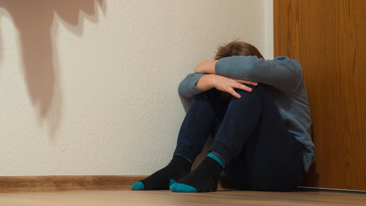 Ein Junge sitzt mit verschränkten Armen vor dem Gesicht in einer Ecke eines Zimmers auf dem Boden.