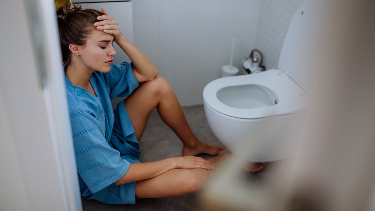 Eine Frau sitzt auf dem Boden vor einer Toilette. Sie hält sich den Kopf und sieht elend aus.