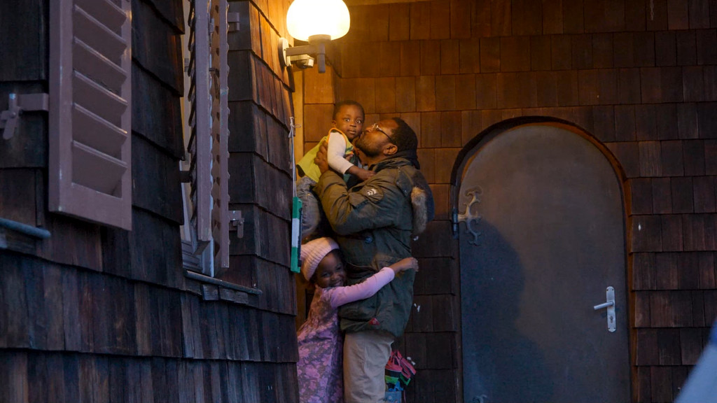 Edward Ebohon hält seine kleine Tochter auf dem Arm während die andere Tochter seine Beine umarmt.