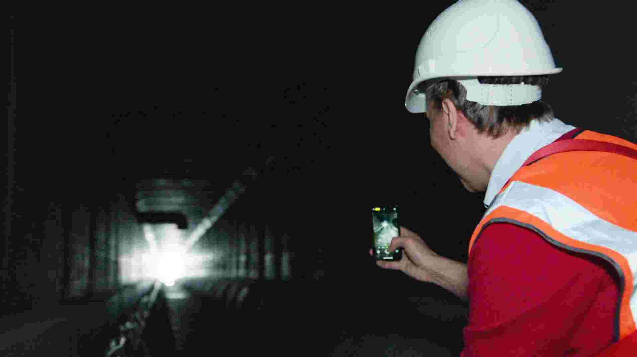 Mann in Schutzkleidung fotografiert mit seinem Handy Wärmetauschermodule in einem Abwasserkanal. Ansicht von hinten.
