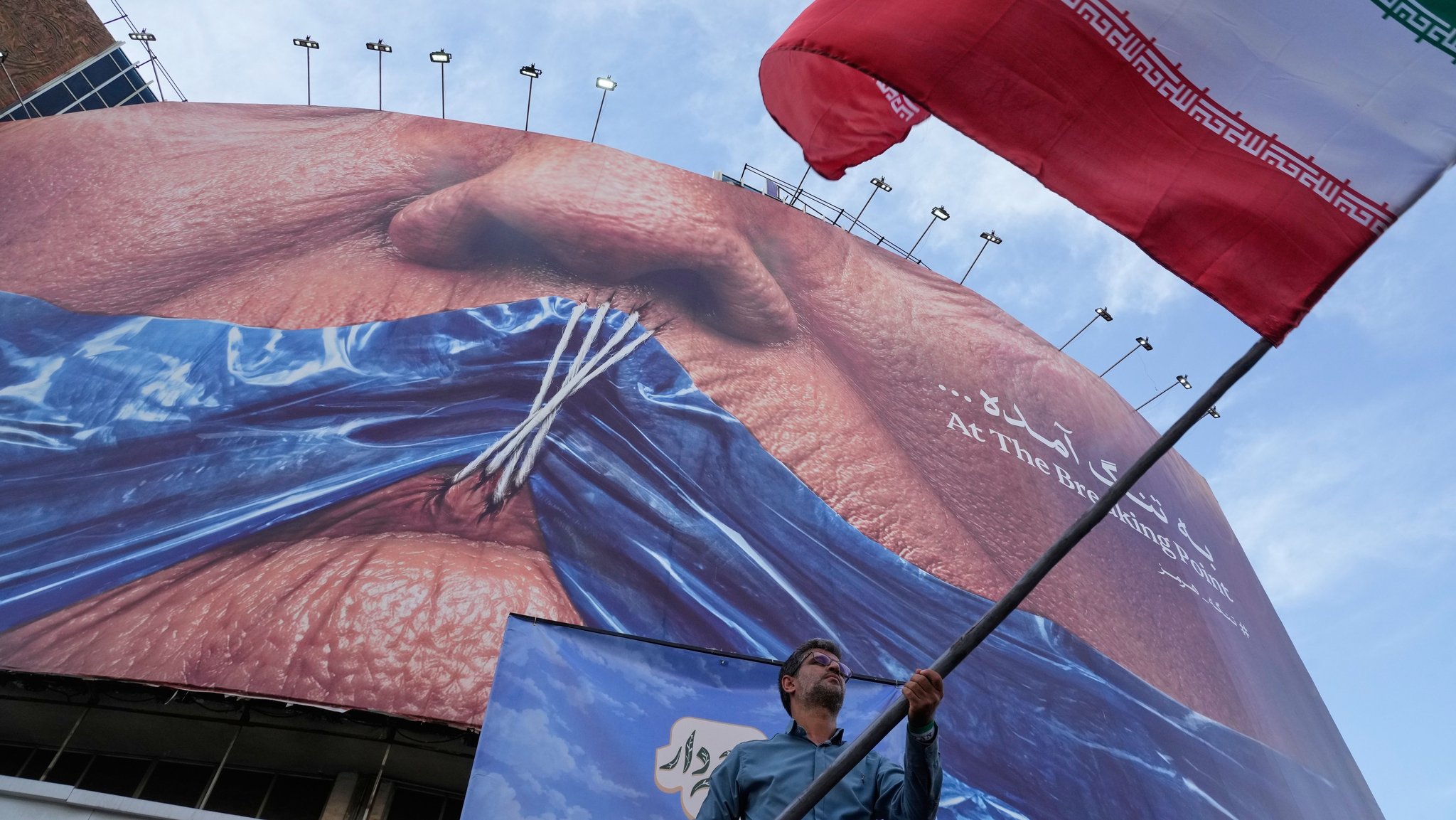 (Symbolbild) Ein Mann schwenkt bei einer regierungsnahen Aktion eine iranische Flagge vor einer Plakatwand mit einer Darstellung der Straße von Hormuz und zugenähten Lippen von US-Präsident Trump. | Bild: dpa-Bildfunk/Vahid Salemi (Symbolbild) Ein Mann schwenkt bei einer regierungsnahen Aktion eine iranische Flagge vor einer Plakatwand mit einer Darstellung der Straße von Hormuz und zugenähten Lippen von US-Präsident Trump.