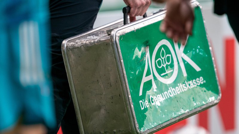 Ein ramponierter Metallkoffer mit der Aufschrift "AOK - Die Gesundheitskasse." wird bei einem Fußballspiel über den Platz getragen.