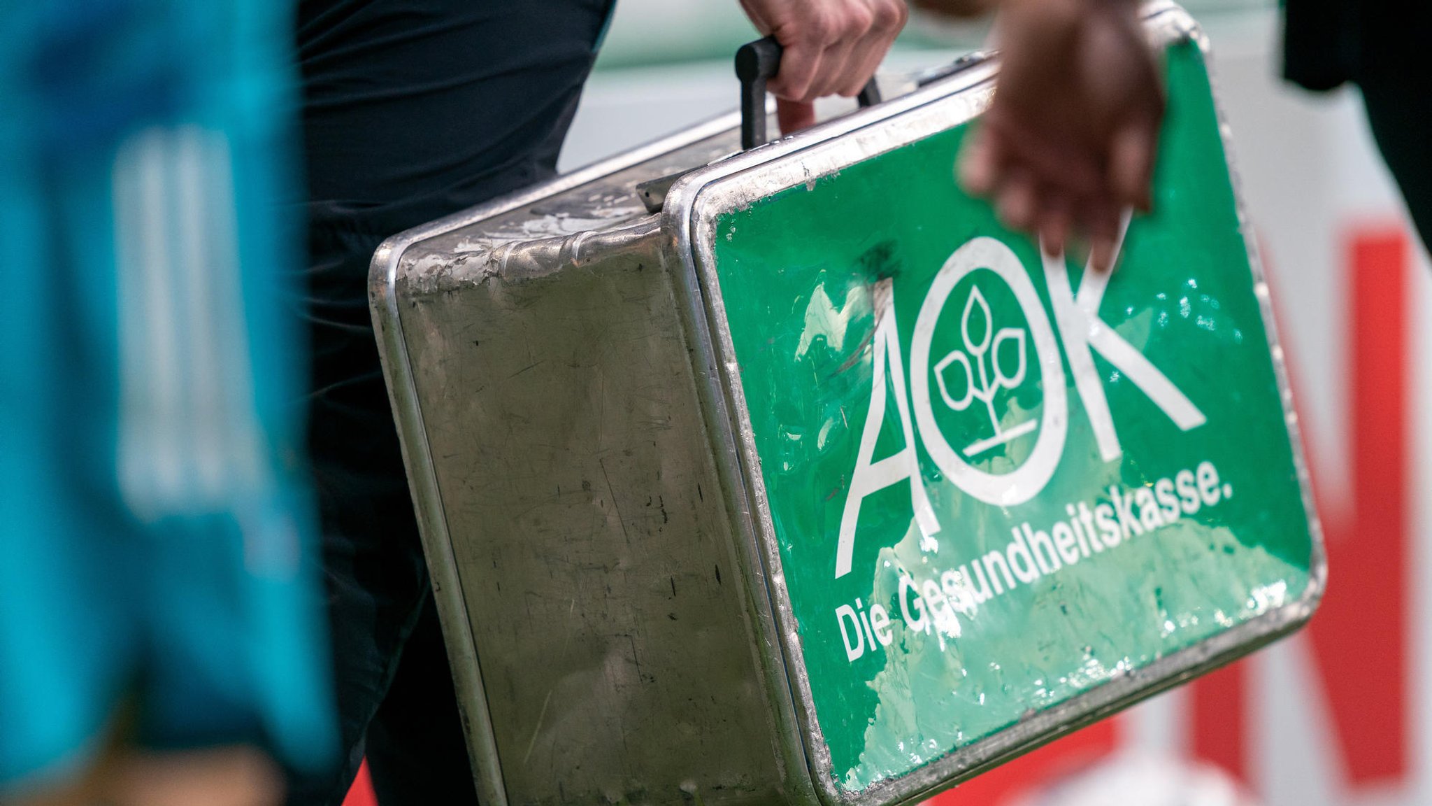 Ein ramponierter Metallkoffer mit der Aufschrift "AOK - Die Gesundheitskasse." wird bei einem Fußballspiel über den Platz getragen.