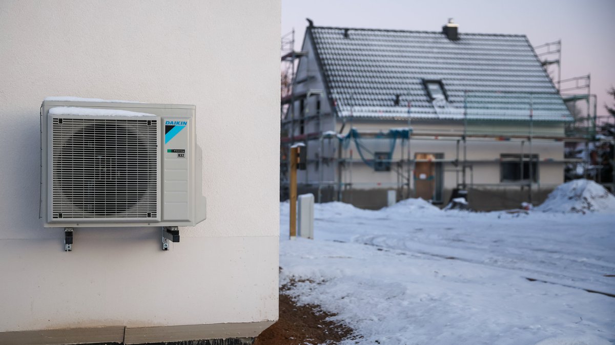 Eine Luftwärmepumpe hängt bei Schnee und Eis an einer Hauswand in einem neu entstehenden Einfamilienhausgebiet.