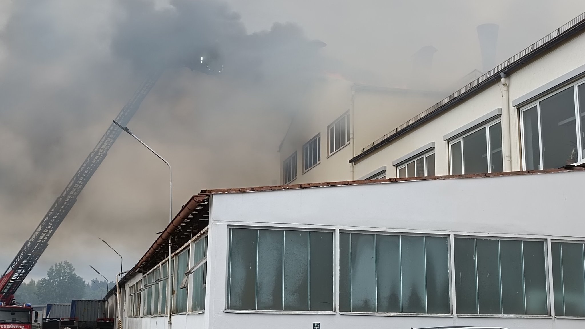 Rauch steigt aus dem Gebäude einer Firma empor. Daneben ist eine Feuerwehrleiter zu sehen.