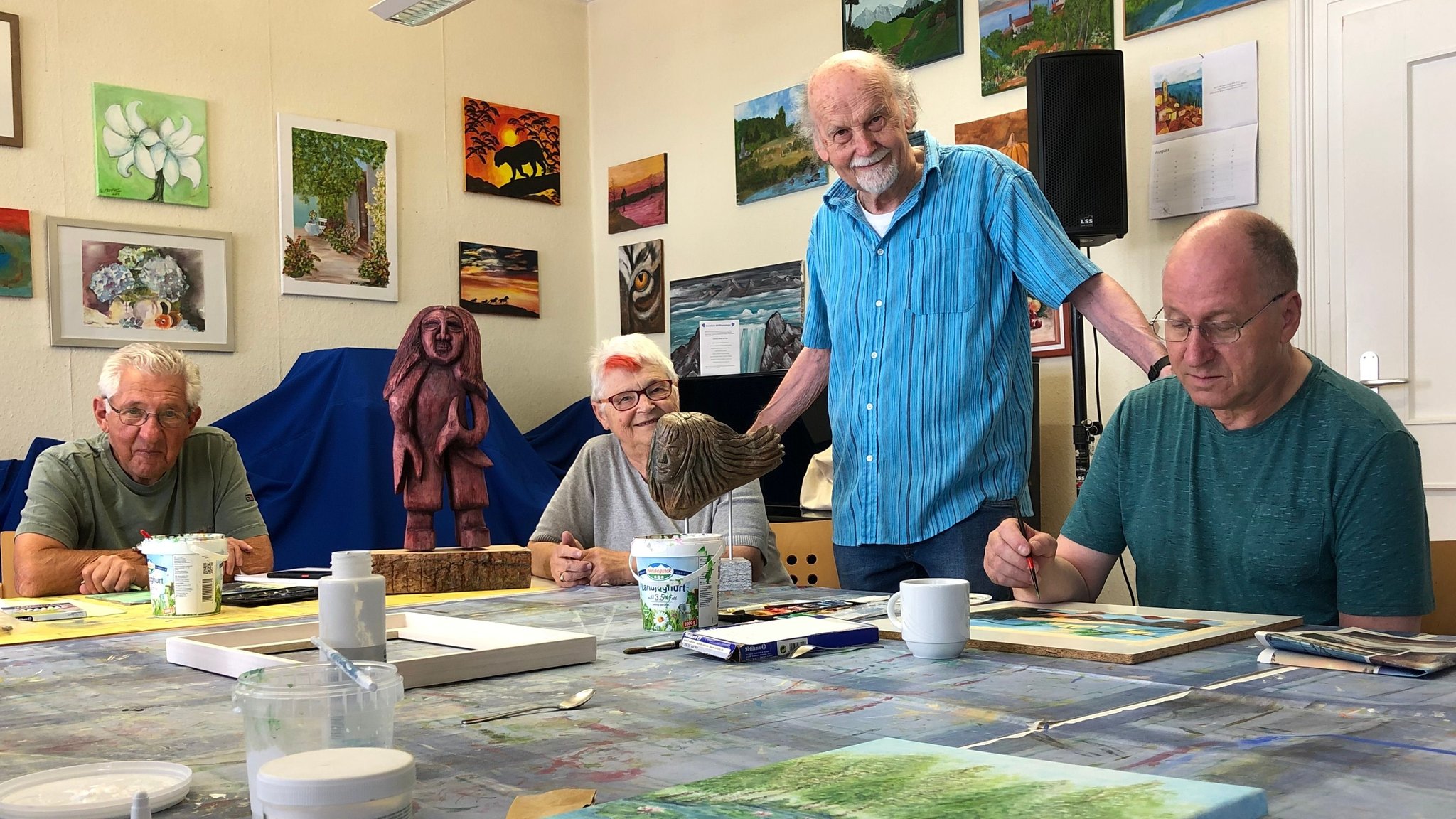 Drei Mitglieder der Kunstgruppe sitzen am Tisch im Kunstraum. In der Mitte steht der Leiter der Gruppe.