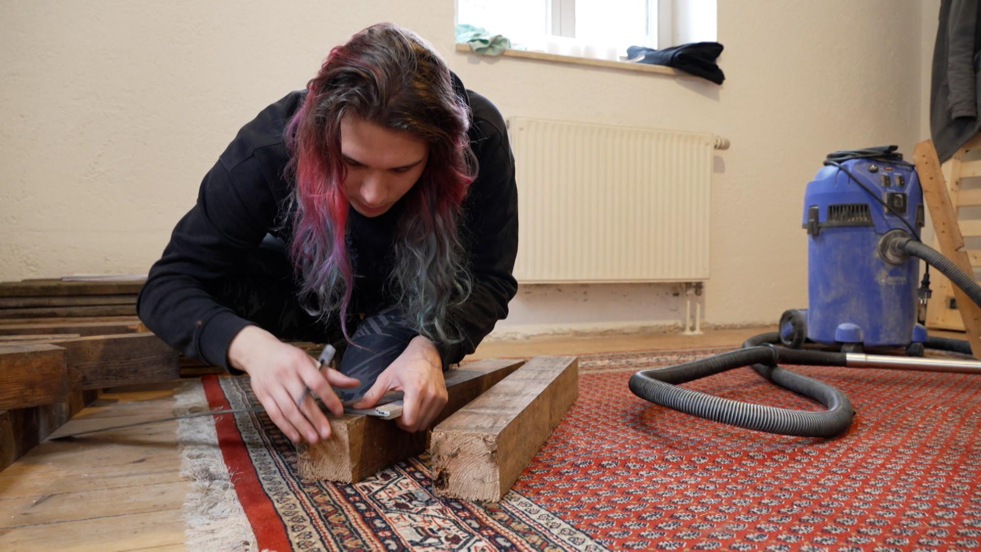 Ein junger Mann mit farbigen Haaren hantiert mit Werkzeug und Holz auf einem ausgelegten Teppich.