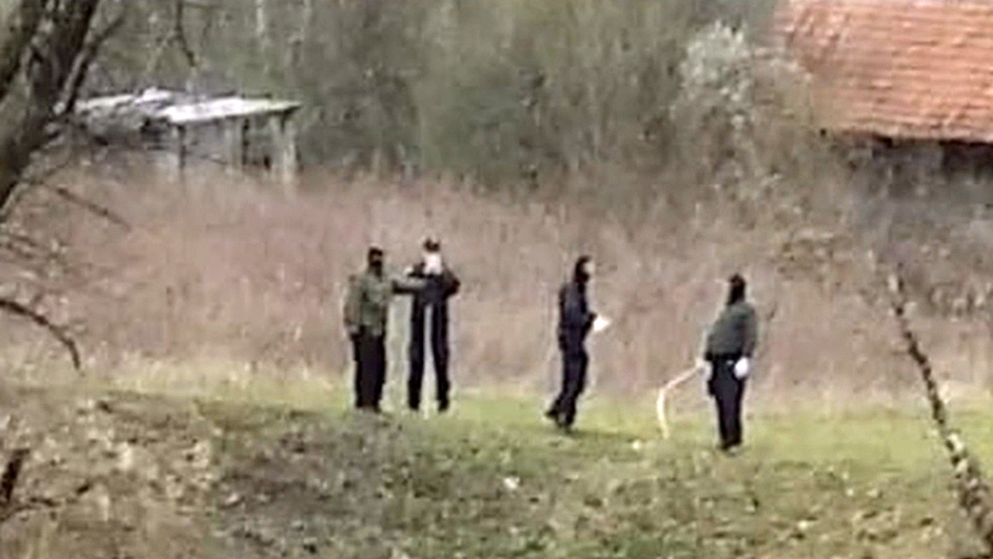 Maskierte Männer in Uniformähnlicher Kleidung stehen an einem Flussufer. Einer hält eine Art Peitsche in der Hand.