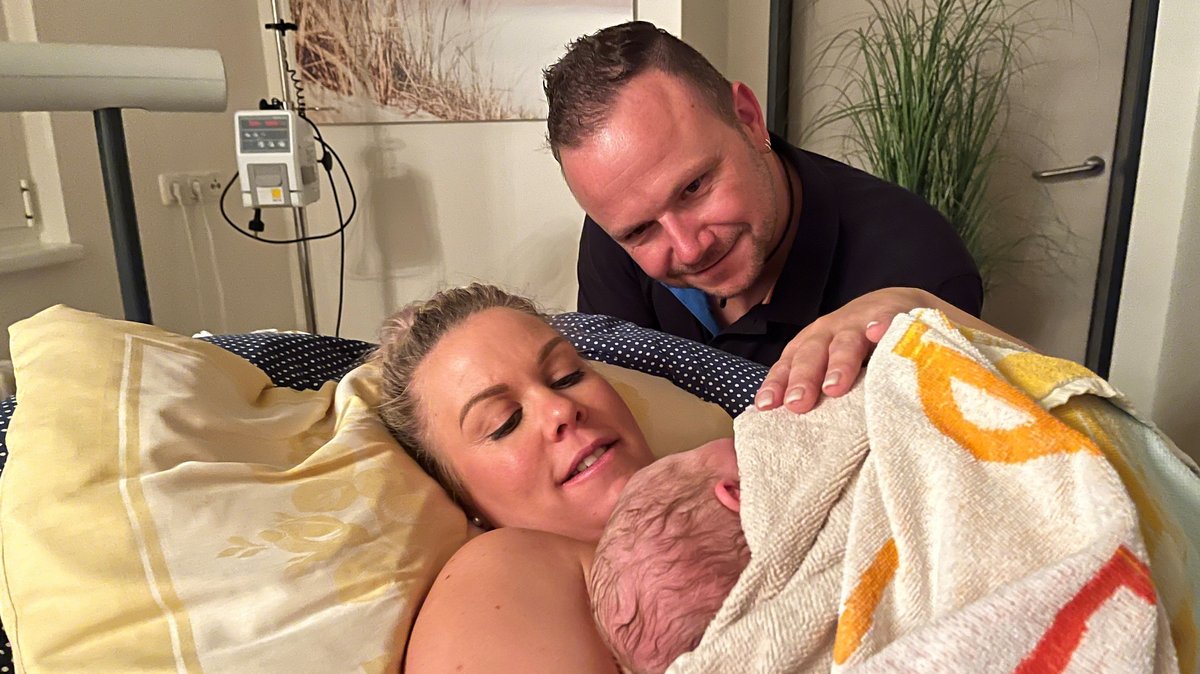 Marita Renner liegt mit ihrem neugeborenen Baby auf der Liege, daneben ihr Mann Andreas Renner - beide lächeln glücklich.