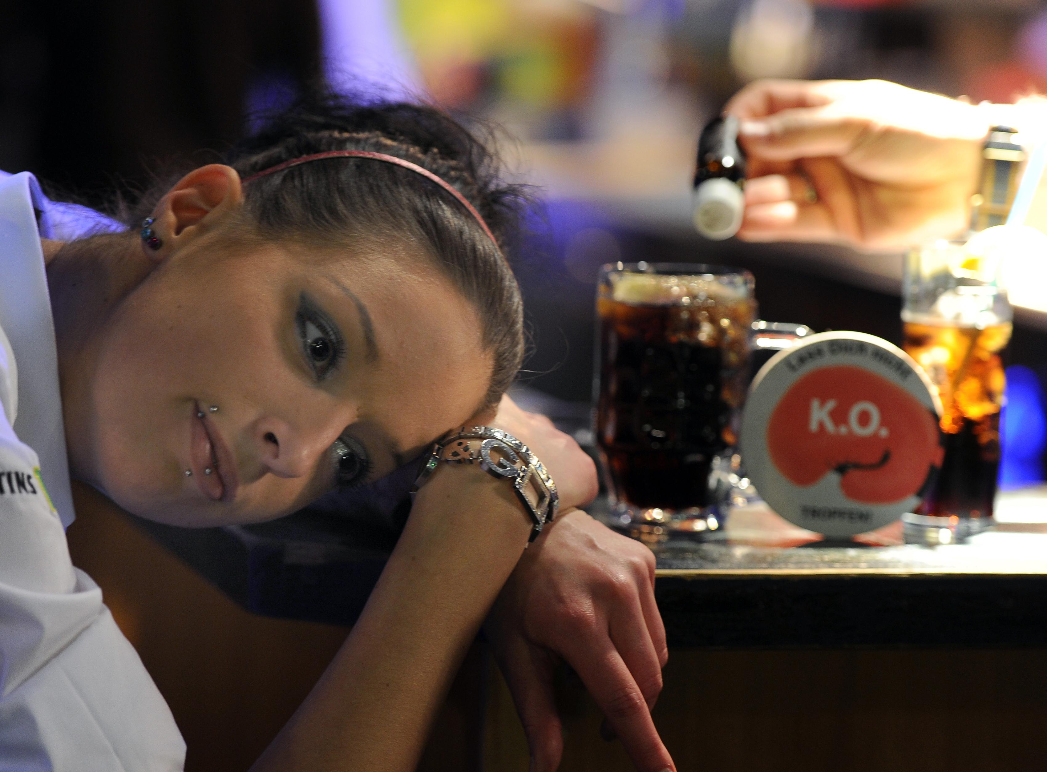 Frau liegt mit dem Kopf auf dem Tisch mit Getränken, eine Hand im Bild füllt K.-o. Tropfen in ein Glas.