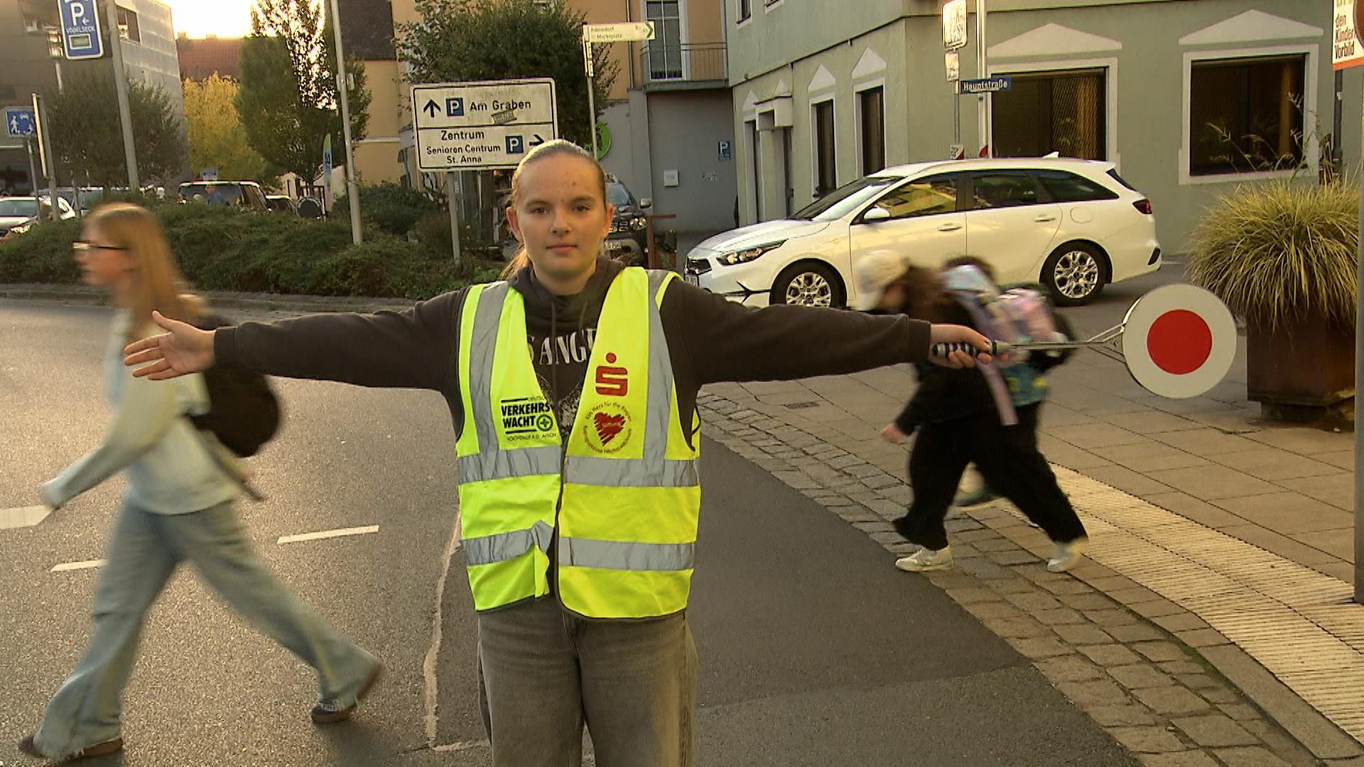 Ein Mädchen steht in Warnweste mir ausgebreiteten Armen auf der Straße - im Hintergrund laufen Schulkinder.