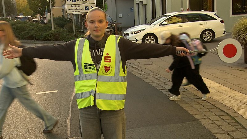 Ein Mädchen steht in Warnweste mir ausgebreiteten Armen auf der Straße - im Hintergrund laufen Schulkinder. | Bild: Bayerischer Rundfunk 2015 Ein Mädchen steht in Warnweste mir ausgebreiteten Armen auf der Straße - im Hintergrund laufen Schulkinder.