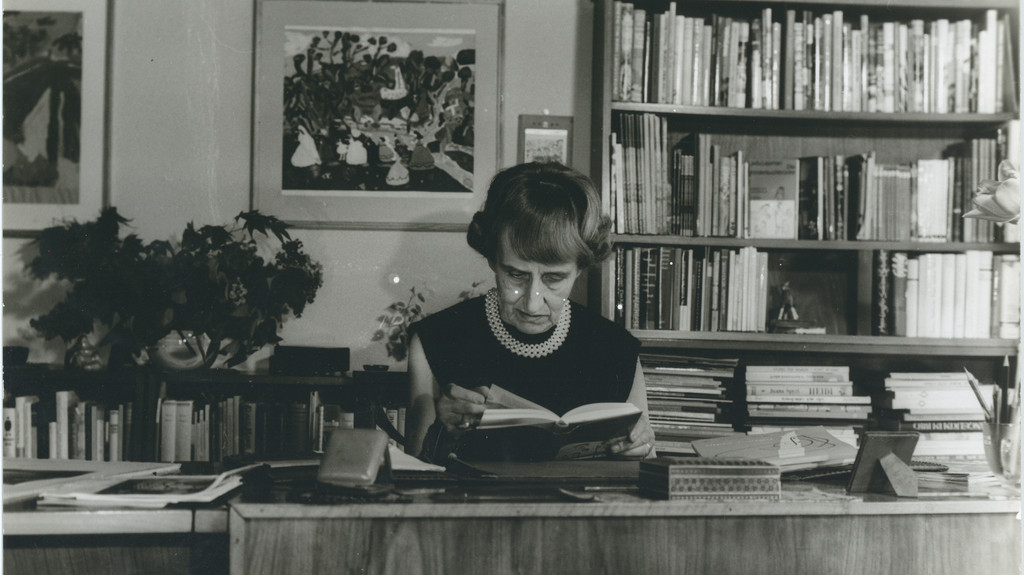 Jella Lepman an ihrem Schreibtisch mit einem Buch in der Hand, hinter ihr ein prall gefülltes Bücherregal