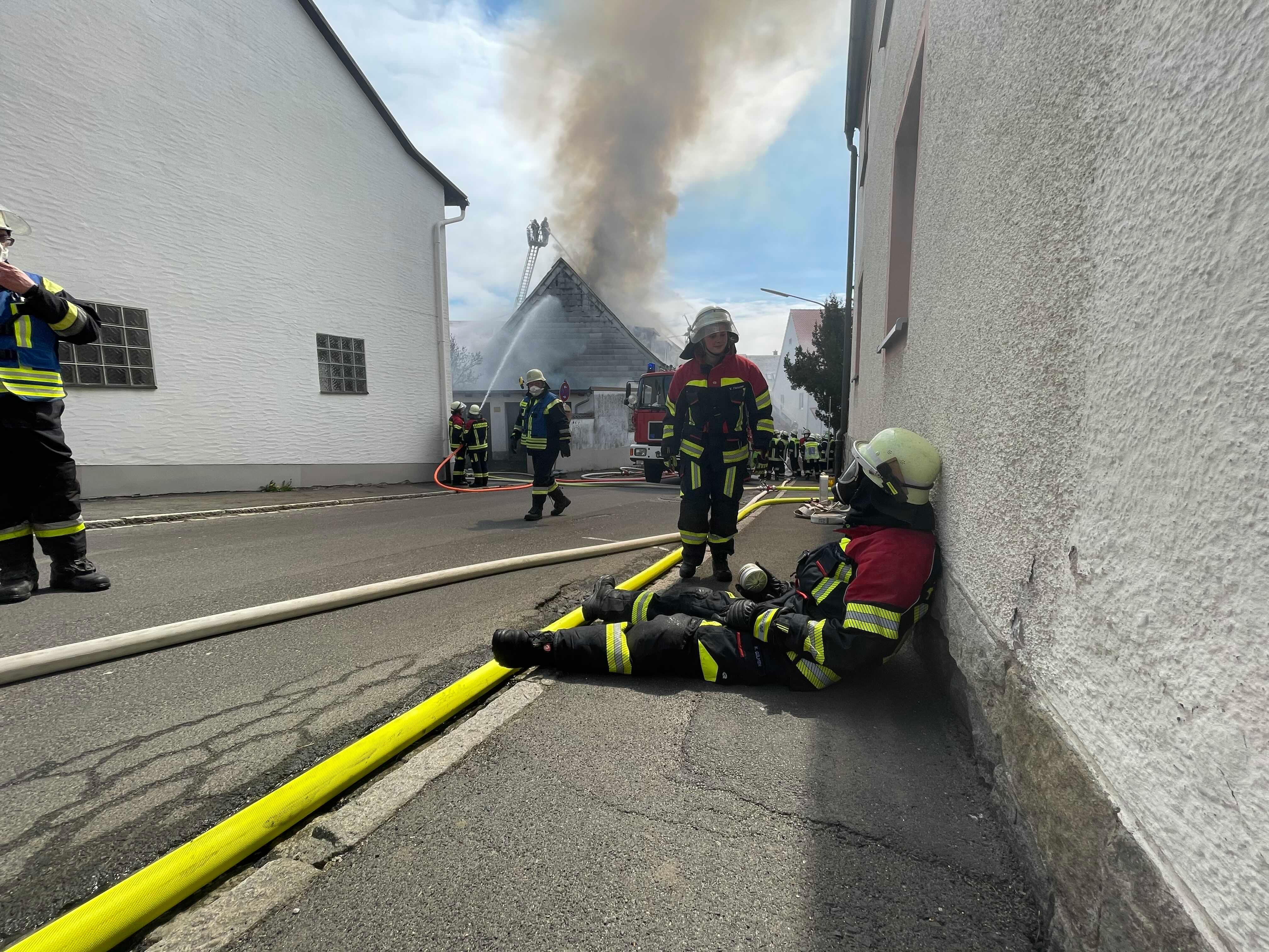 Ein Feuerwehrmann sitzt am Boden, im Hintergrund tritt Rauch von einem brennenden Gebäude auf. | Bild:News5/Fricke