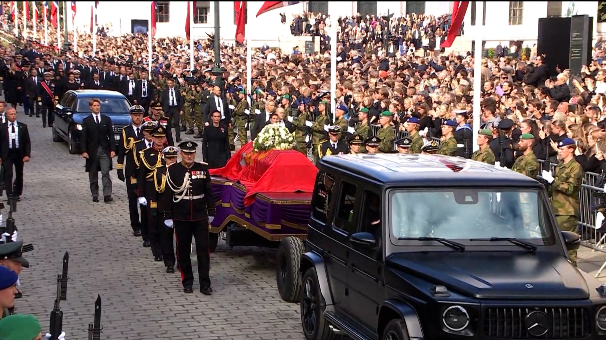 Ein schwarzes Auto mit dem Sarg Haralds V. von Norwegen, über den ein knallrotes Tuch gebreitet ist, fährt durch eine Menschenmenge und ein Spalier von Soldaten. | Bild: Bayerischer Rundfunk 2026 Ein schwarzes Auto mit dem Sarg Haralds V. von Norwegen, über den ein knallrotes Tuch gebreitet ist, fährt durch eine Menschenmenge und ein Spalier von Soldaten.