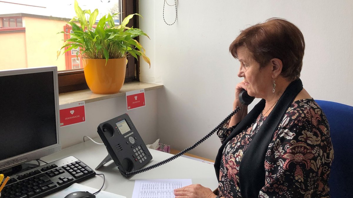 Eine Frau sitzt an einem Schreibtisch in einem Büro und telefoniert. Vor ihr liegt auf dem Schreibtisch eine Liste mit Telefonnummern und Namen. Sie trägt eine Bluse und einen schwarzen Schal.
