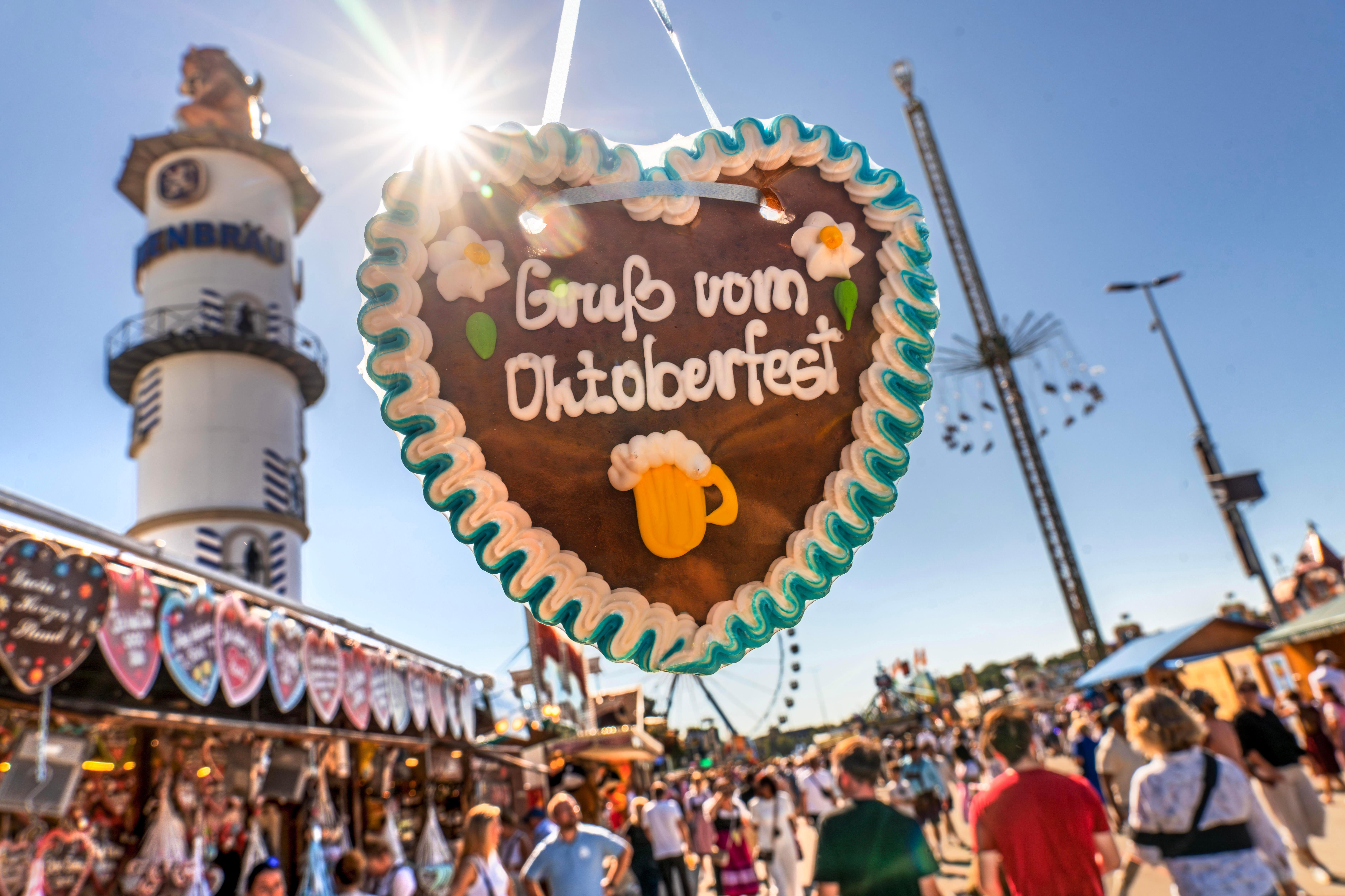 Nahaufnahme eines Lebkuchenherzes mit der Aufschrift "Gruß vom Oktoberfest" im Gegenlicht, dahinter Menschenmengen und Fahrgeschäfte
