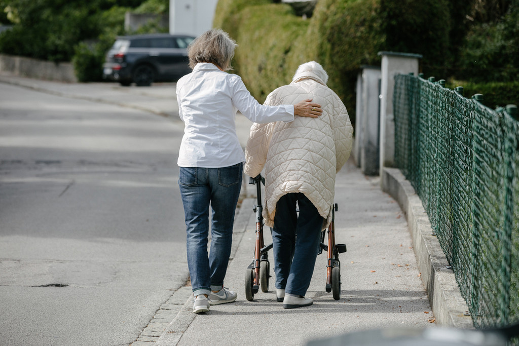 Eine Frau stützt eine Seniorin, die an einem Rollator läuft.
